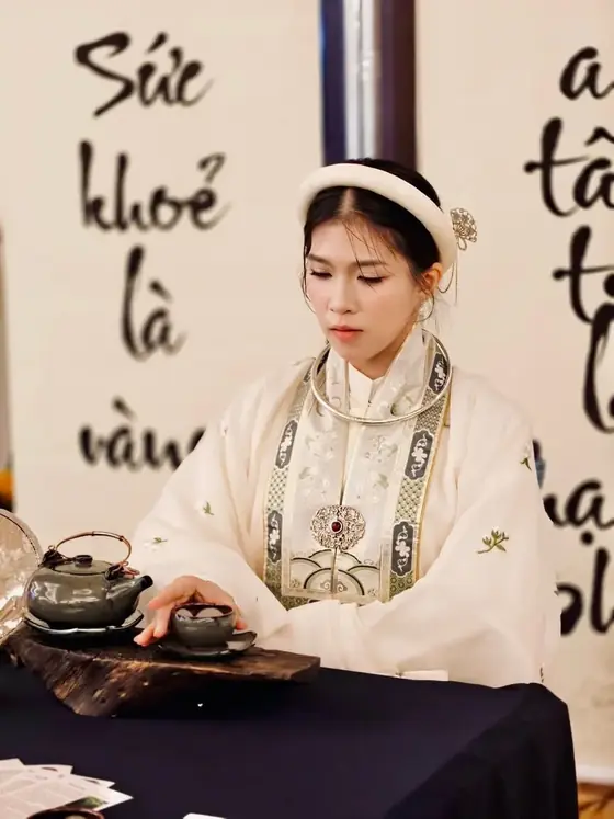 Vy Đan bên bàn trà trước thư pháp Sức khỏe là vàng